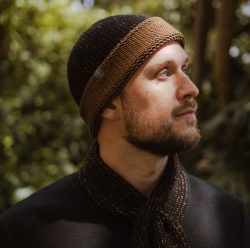 A model standing outdoors wearing a two tone Marbled Cuffed Toque in Oxen. The Toque features a silver bean at the crown. This is shown here with a matching Ascot Scarf.