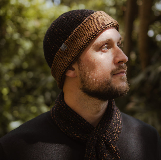 A model standing outdoors wearing a two tone Marbled Cuffed Toque in Oxen. The Toque features a silver bean at the crown. This is shown here with a matching Ascot Scarf.