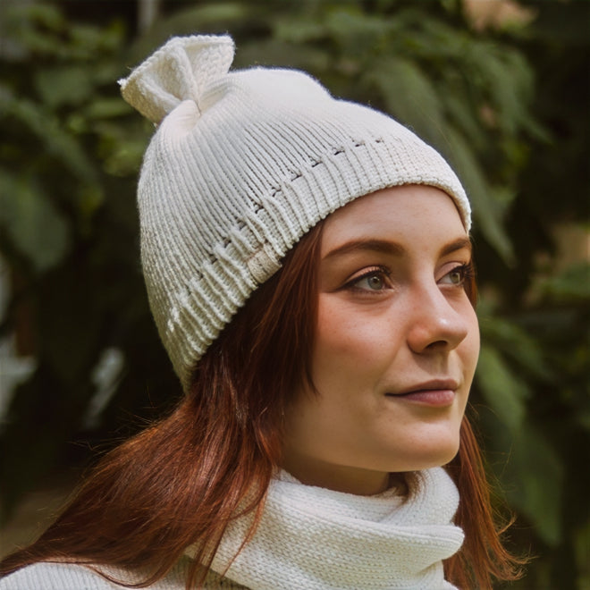 A model wearing a french vanilla -colored merino wool beanie with a distinctive 'petal gather' at the crown, paired with a matching infinity scarf.
